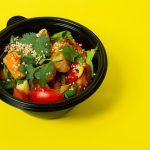From above of delicious healthy salad consisting of various vegetables in plastic bowl placed on bright yellow background